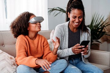 Happy mother using smart phone sitting on sofa with daughter wearing virtual reality simulator at home