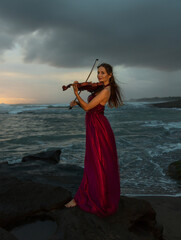 Beautiful Caucasian woman with violin on the beach. Music and art concept. Slim girl wearing long red dress and playing violin in nature. Sunset time. Cloudy sky. Bali © Olga