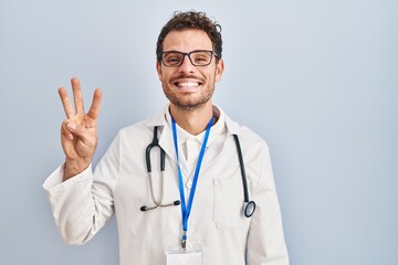 Fototapeta premium Young hispanic man wearing doctor uniform and stethoscope showing and pointing up with fingers number three while smiling confident and happy.