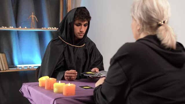 Man witch sits at table in dark gothic clothes. Art costume. Fortune teller magical man reading future on tarot cards for client. Ritual candles burning, divination
