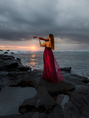 Caucasian woman with violin on the beach. Music and art concept. Slim girl wearing long red dress and playing violin in nature. Sunset time. Cloudy sky. View from back. Bali