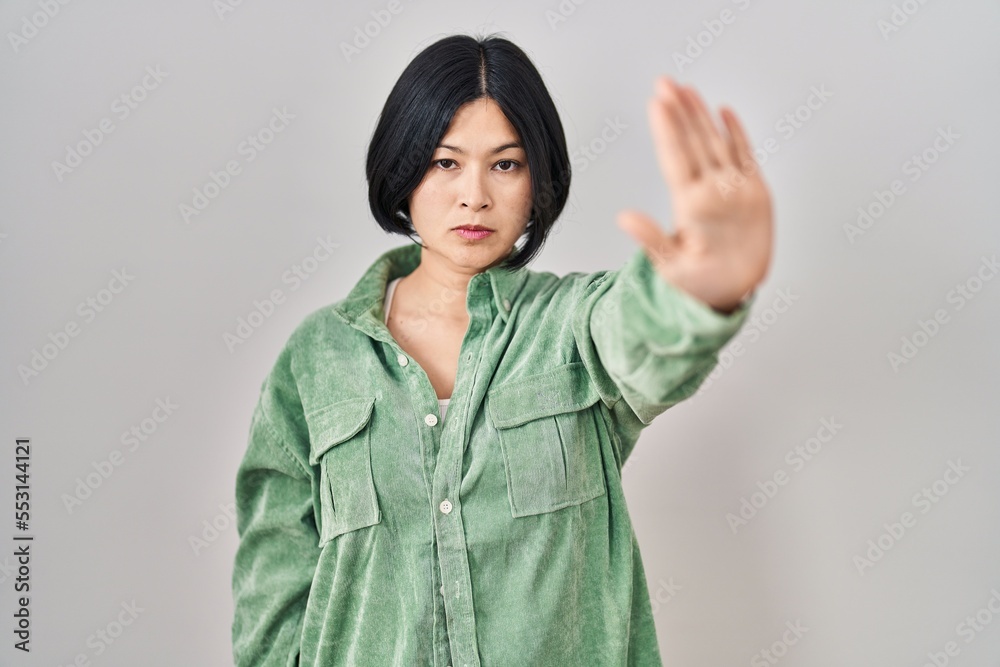 Sticker Young asian woman standing over white background doing stop sing with palm of the hand. warning expression with negative and serious gesture on the face.