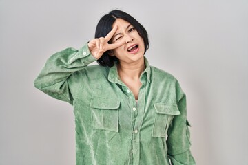 Young asian woman standing over white background doing peace symbol with fingers over face, smiling cheerful showing victory