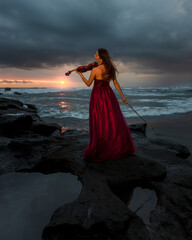 Caucasian woman with violin on the beach. Music and art concept. Slim girl wearing long red dress and playing violin in nature. Sunset time. Cloudy sky. View from back. Bali