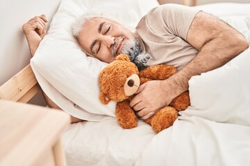 Obraz na płótnie Canvas Middle age grey-haired man hugging teddy bear lying on bed sleeping at bedroom