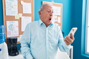 Senior man with grey hair working at the office doing video call with smartphone scared and amazed with open mouth for surprise, disbelief face