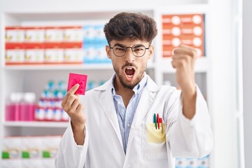 Arab man with beard working at pharmacy drugstore holding condom afraid and shocked with surprise and amazed expression, fear and excited face.