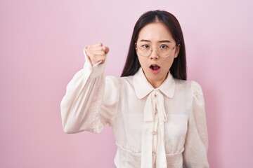 Young chinese woman standing over pink background angry and mad raising fist frustrated and furious...