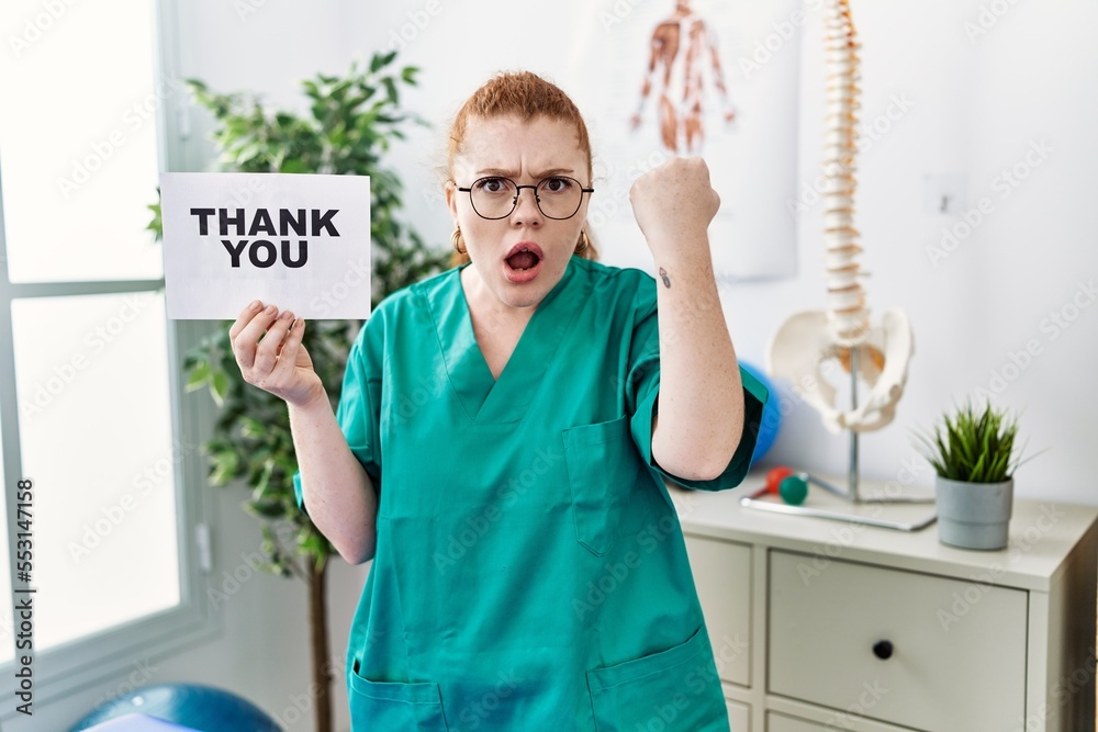 Wall mural young redhead physiotherapist woman working at pain recovery clinic holding thank you banner annoyed