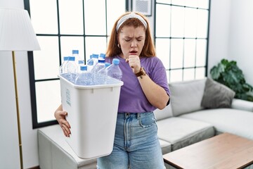 Young redhead woman holding recycling wastebasket with plastic bottles feeling unwell and coughing...