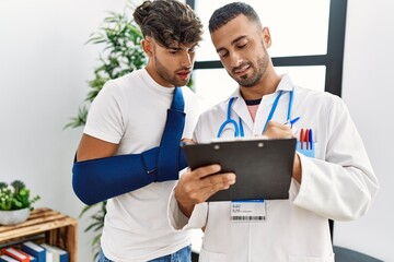 Two hispanic men doctor and patient writing on clipboard standing at hospital waiting room