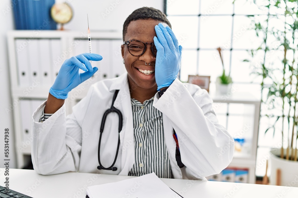 Poster young african doctor man holding syringe at the hospital covering one eye with hand, confident smile