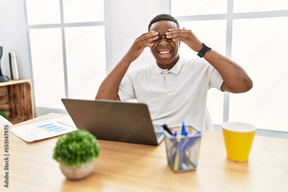 Poster young african man working at the office using computer laptop covering eyes with hands smiling cheer