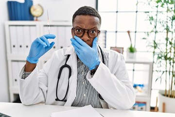 Young african doctor man holding syringe at the hospital shocked covering mouth with hands for mistake. secret concept.