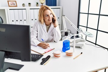 Young blonde woman wearing dentist uniform speaking on the smartphone at dental clinic