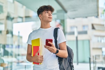 Young hispanic teenager student using smartphone holding books at university