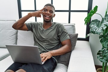 Young african american man using laptop at home sitting on the sofa doing peace symbol with fingers over face, smiling cheerful showing victory