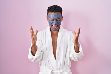 Young hispanic man wearing beauty face mask and bath robe celebrating mad and crazy for success...