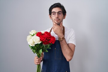 Young hispanic man holding bouquet of white and red roses looking confident at the camera smiling with crossed arms and hand raised on chin. thinking positive.