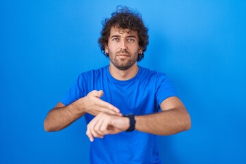 Hispanic young man standing over blue background in hurry pointing to watch time, impatience, upset and angry for deadline delay