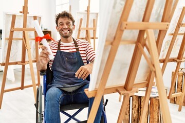 Young hispanic man holing art diploma sitting on wheelchair at art studio