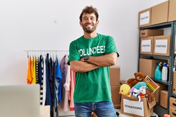 Young hispanic volunteer man smiling happy standing with arms crossed gesture at charity center.