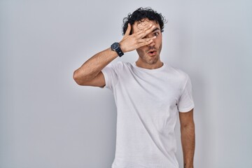 Hispanic man standing over isolated background peeking in shock covering face and eyes with hand,...