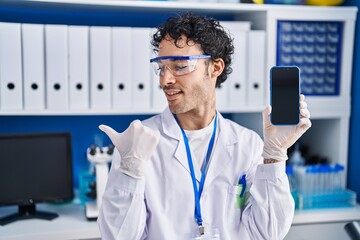 Hispanic man working at scientist laboratory showing smartphone screen pointing thumb up to the side smiling happy with open mouth