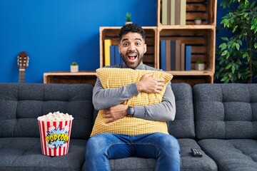 Hispanic man with beard eating popcorn watching a movie at home celebrating crazy and amazed for...