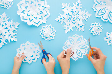 Adult mother and child hands cutting different white snowflake shapes from paper on light blue...