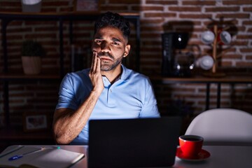 Fototapeta na wymiar Hispanic man with beard using laptop at night touching mouth with hand with painful expression because of toothache or dental illness on teeth. dentist