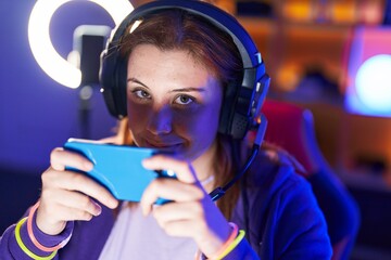 Young beautiful plus size woman streamer playing video game using smartphone at gaming room