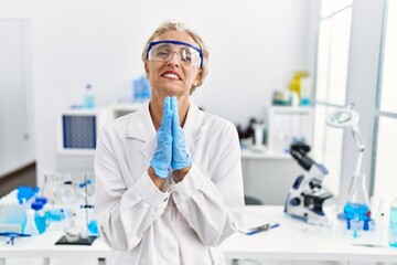 Middle age blonde woman working at scientist laboratory begging and praying with hands together with hope expression on face very emotional and worried. begging.