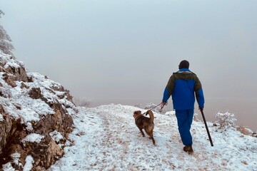 Man and dog on a snowy mountain path with dense fog in the background. - 553151541