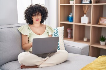 Fototapeta na wymiar Young brunette woman with curly hair using laptop sitting on the sofa at home skeptic and nervous, frowning upset because of problem. negative person.