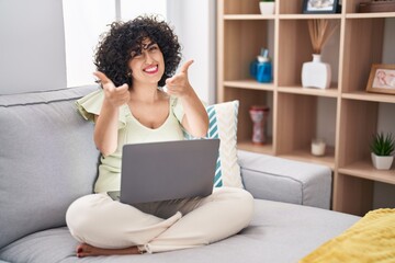 Young brunette woman with curly hair using laptop sitting on the sofa at home pointing fingers to camera with happy and funny face. good energy and vibes.