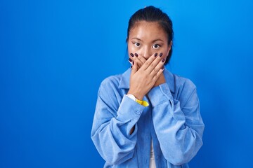 Asian young woman standing over blue background shocked covering mouth with hands for mistake....