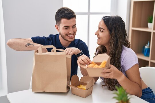 Young Hispanic Couple Eating Take Away Food Sitting On Table At Home