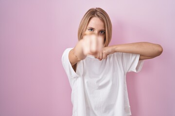 Young caucasian woman standing over pink background punching fist to fight, aggressive and angry...