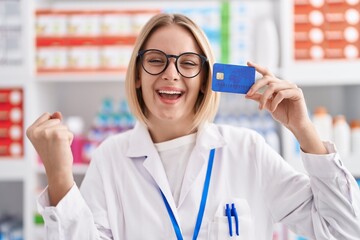 Young caucasian woman working at pharmacy drugstore holding credit card screaming proud, celebrating victory and success very excited with raised arm