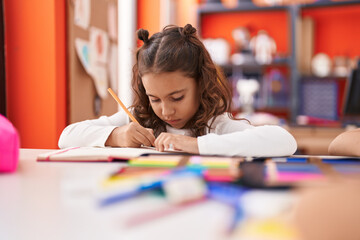 Adorable hispanic girl student sitting on table writing on notebook at classroom