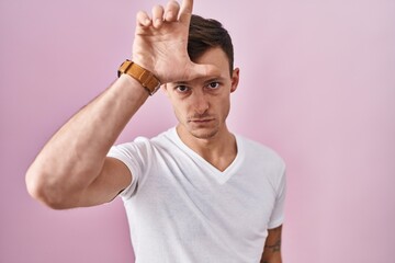 Caucasian man standing over pink background making fun of people with fingers on forehead doing loser gesture mocking and insulting.