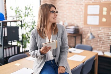 Young hispanic woman working at the office wearing glasses looking away to side with smile on face,...