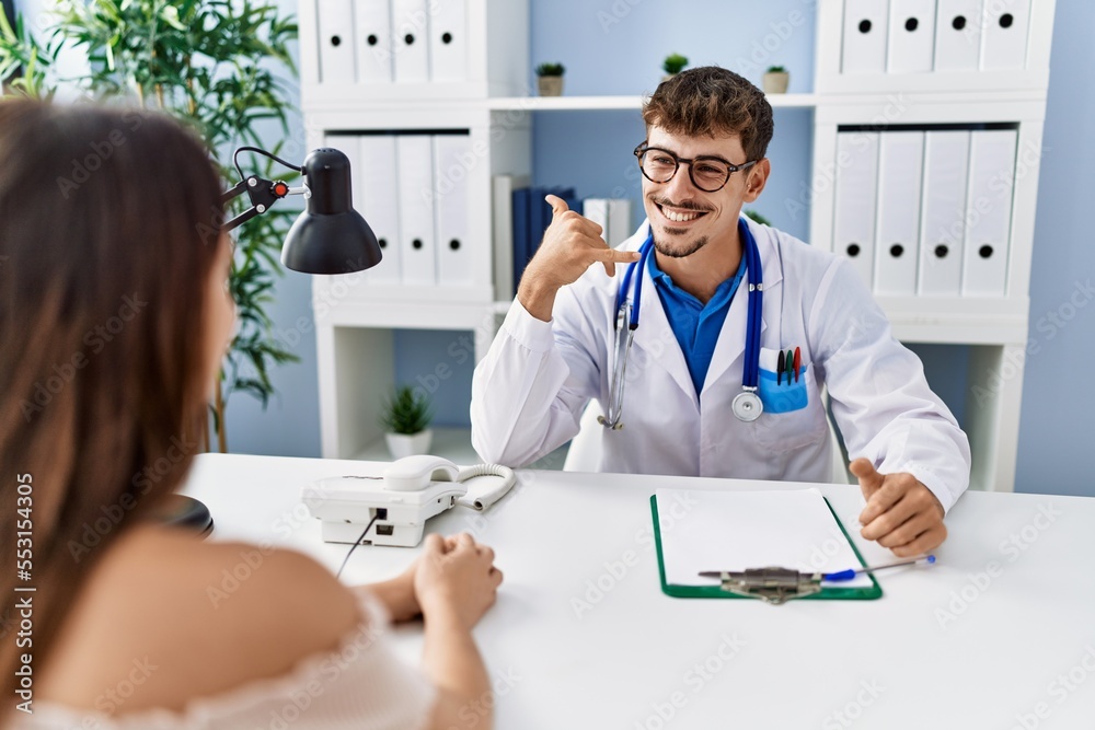 Sticker young doctor with client at medical clinic smiling doing phone gesture with hand and fingers like ta