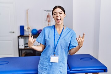 Young hispanic woman wearing physiotherapist uniform standing at clinic shouting with crazy...