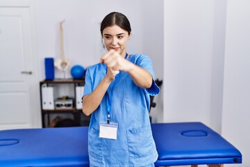Young hispanic woman wearing physiotherapist uniform standing at clinic punching fist to fight,...