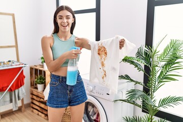 Young hispanic woman cleaning dirty white t shirt with stain at laundry room winking looking at the...
