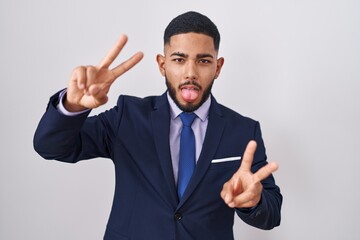 Young hispanic man wearing business suit and tie smiling with tongue out showing fingers of both hands doing victory sign. number two.