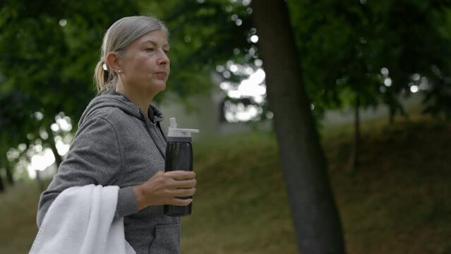 Mature woman drinking water from the sport bottle while walking in the park after training. Elderly female feeling thirsty. Aging youthfully.