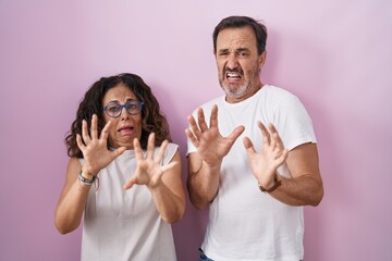 Middle age hispanic couple together over pink background afraid and terrified with fear expression stop gesture with hands, shouting in shock. panic concept.